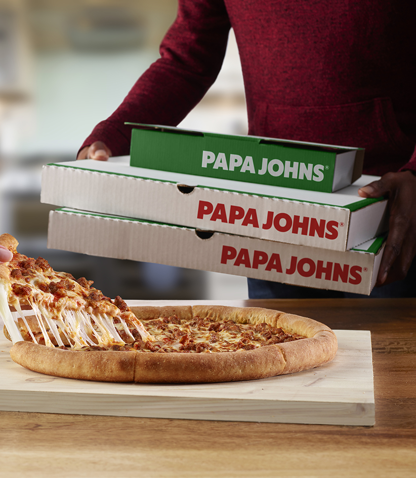 Man in a red sweater holding three Papa John's boxes and on the table shown a female's hand taking a slice Papa Johns' epic stuffed crust sausage pizza. Kitchen setting, also shown stack of plates with a garlic dip and a pepperoncini.
